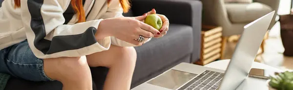 In a cozy setting, a young beautiful woman relaxes with a laptop and a green apple in hand. — Stock Photo