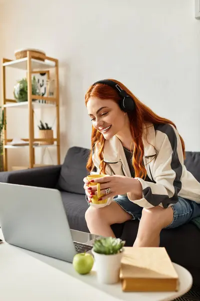 A cheerful young woman relaxes on a couch, sipping a beverage while engaged with her laptop. — стоковое фото