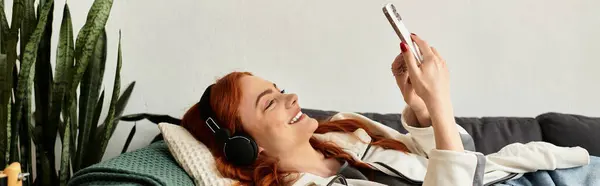 A young woman with stunning red hair smiles while enjoying music on her phone. — Stock Photo
