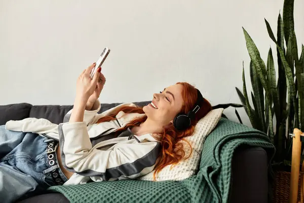 Una giovane bella donna si sdraia su un morbido divano con le cuffie, sorridendo mentre usa il suo telefono. — Foto stock