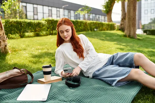 A young woman relaxes on a green blanket, sipping coffee and scrolling her phone in the park. — Stock Photo