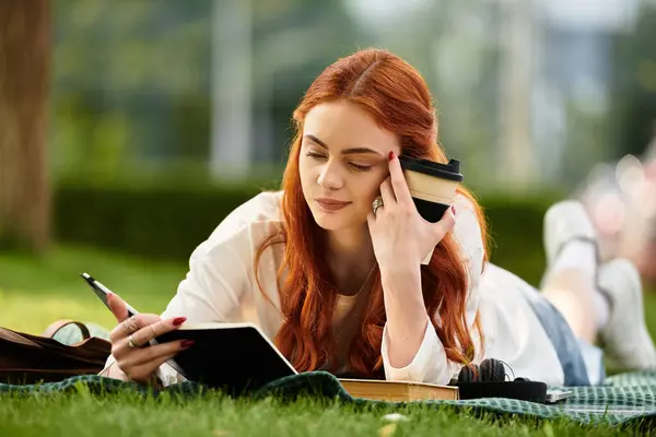 Una giovane bella donna giace sull'erba, scrive in un diario e assapora il suo caffè nel parco.. — Foto stock
