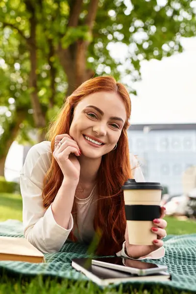 Eine junge schöne Frau entspannt sich im Gras, nippt an ihrem Kaffee und lächelt strahlend. — Stockfoto