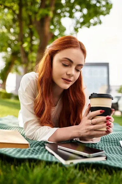 Una joven hermosa mujer disfruta de su café mientras está acostada sobre una manta en un parque soleado. - foto de stock