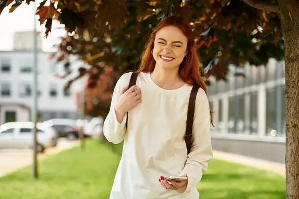 A joyful young woman with red hair smiles while standing on a sunny day in a vibrant green park. — Stock Photo