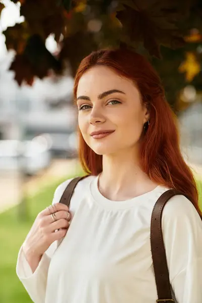 Young woman with red hair stands under autumn foliage, radiating warmth and charm. — Stock Photo