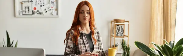 Relaxed young woman with red hair sips a warm drink while checking her phone indoors. — Stock Photo