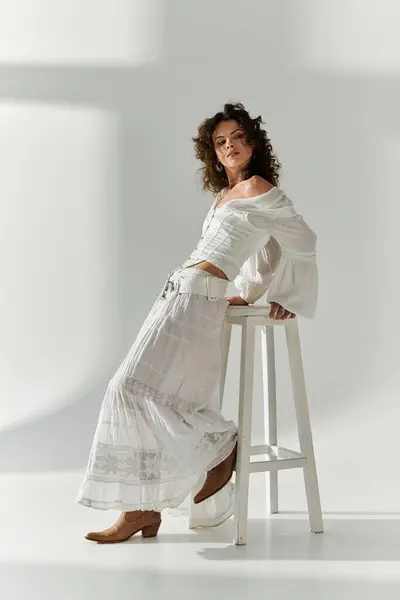 A stylish young woman in white leans on a stool, highlighting her chic outfit in soft light. — Stock Photo