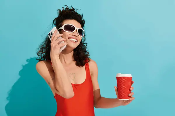 A stylish woman in a red outfit joyfully drinks coffee while making a call. — стоковое фото