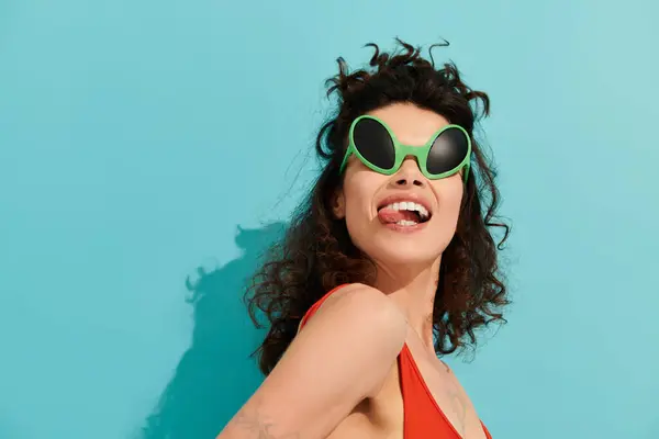 Una mujer con estilo posa juguetonamente sobre un fondo azul brillante, haciendo alarde de sus gafas de sol frescas. - foto de stock