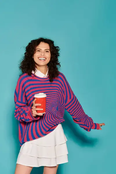 Brightly dressed young woman smiles cheerfully, holding a coffee cup against a turquoise backdrop. — Stock Photo