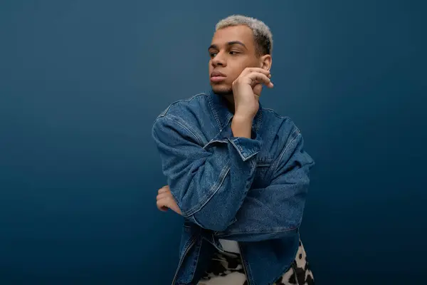 Un giovane uomo sicuro di sé posa con stile, mostrando la sua giacca di jeans alla moda in mezzo a uno sfondo blu. — Foto stock