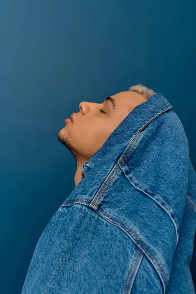 A young Black man shows off his dyed hair and style in a denim jacket by a blue wall. — Foto stock