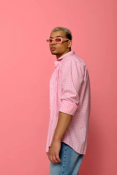 Confident young man showcases his unique style with sunglasses and a checkered shirt in pink. — Stock Photo