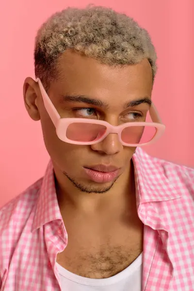 Un joven hombre negro muestra su estilo de moda con el pelo teñido y gafas de sol en un telón de fondo rosa. - foto de stock