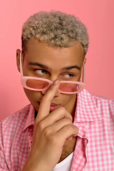 A young fashionable man with dyed hair poses playfully in a checkered shirt and pink shades. — Stock Photo