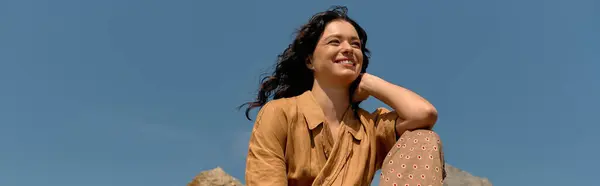 A young woman with curly hair beams while dressed in a trendy summer outfit against a clear sky, banner — стоковое фото