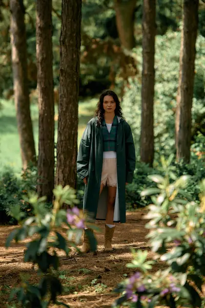 A young woman in a stylish raincoat walks through a lush green forest, enjoying nature. — Stock Photo