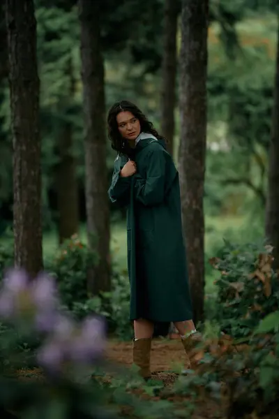 Stylish young woman with curly hair explores a vibrant green forest while dressed in a raincoat. — Stock Photo