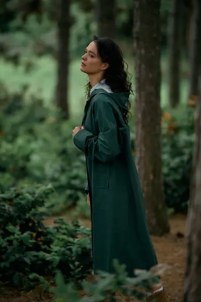 Curly-haired woman stands gracefully in a vibrant green forest wearing a trendy raincoat. — стоковое фото