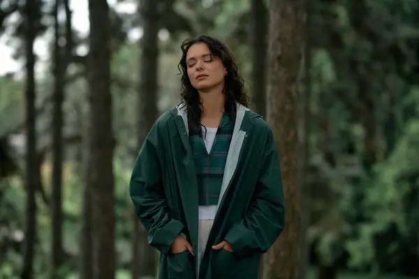 A young woman in a stylish raincoat stands among tall trees, embracing the tranquility of nature. — стоковое фото