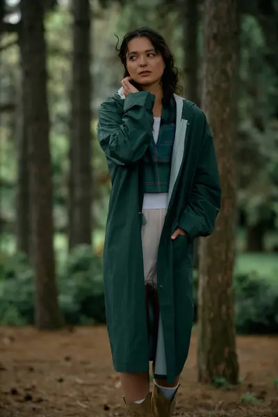 En un exuberante bosque de verano, una joven con el pelo rizado lleva un impermeable elegante mientras refleja. - foto de stock