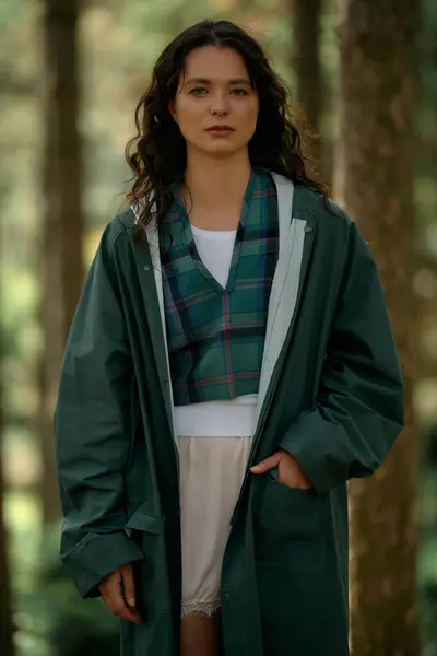 A young woman with curly hair stands confidently in a vibrant forest while wearing a raincoat. — Fotografia de Stock