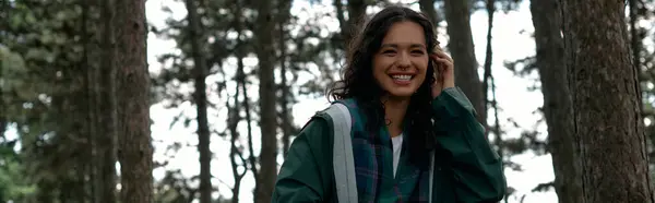 A young woman with curly hair is smiling while exploring a vibrant forest wearing a trendy raincoat, banner — Stock Photo