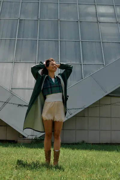 With her curly hair flowing, a young woman poses joyfully in a trendy raincoat amidst green grass. — стоковое фото