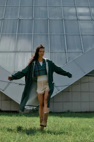A young woman with curly hair strolls gracefully outdoors in a trendy raincoat and shorts. — стоковое фото