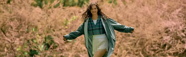A young woman with curly hair poses in a chic raincoat, enjoying a vibrant outdoor setting, banner — Stock Photo