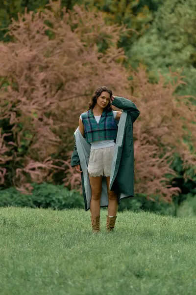 A young woman sporting a fashionable raincoat poses amidst vibrant greenery and flowers. — Stock Photo