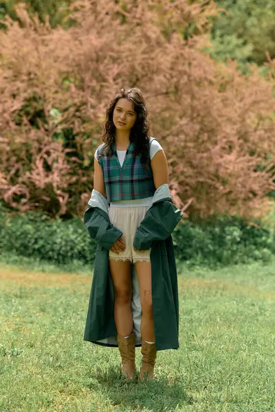 A young woman with curly hair playfully poses in a fashionable raincoat surrounded by nature. — стоковое фото