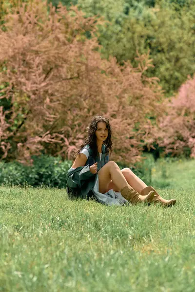 A young woman with curly hair poses in a fashionable raincoat, surrounded by blooming foliage. — Stock Photo