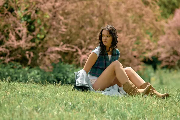 Curly-haired woman poses gracefully in a stylish raincoat on lush green grass surrounded by flowers. — Stock Photo