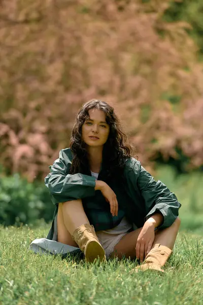 Young woman with curly hair sits comfortably on the grass, showcasing her trendy outfit in summer. — стокове фото