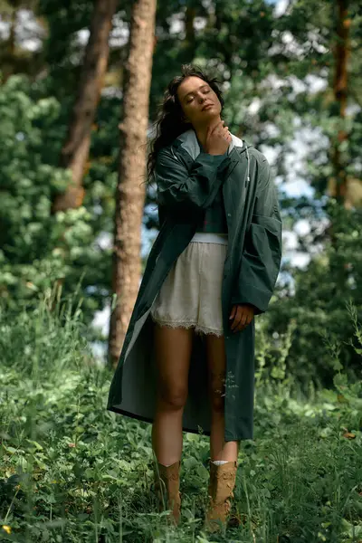 A young woman with curly hair poses gracefully outdoors in a fashionable raincoat amidst nature. — Stock Photo