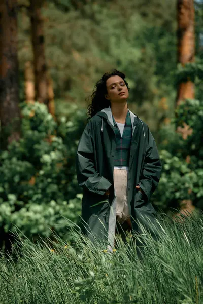 Young woman with curly hair stands confidently in a stylish raincoat amidst lush greenery. — Stock Photo