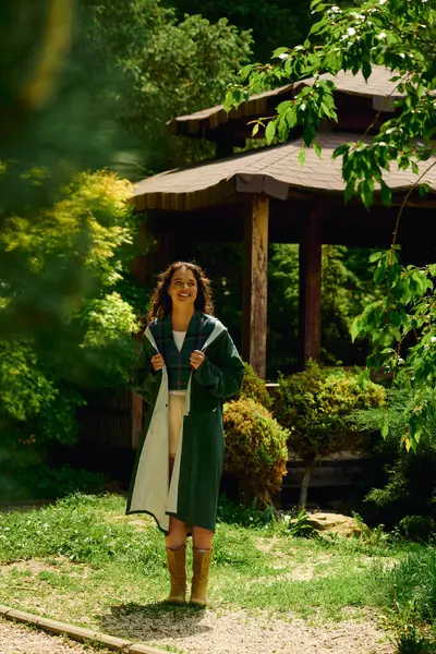 A young woman in a trendy raincoat stands proudly in sunlight among lush greenery. — стоковое фото