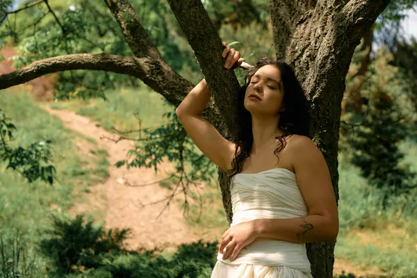 A young woman with curly hair stands gracefully by a tree, embracing the warmth of summer. — Stock Photo