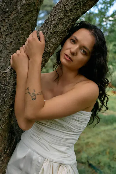 A young woman in a white dress stands gracefully by a tree on a summer afternoon. — Stock Photo