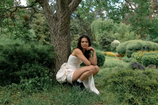 A young woman gracefully poses in a garden, wearing a white dress that matches the greenery. — стоковое фото