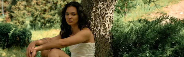 A young woman with curly hair in white, posed amid lush greenery on a warm summer day, banner — стоковое фото