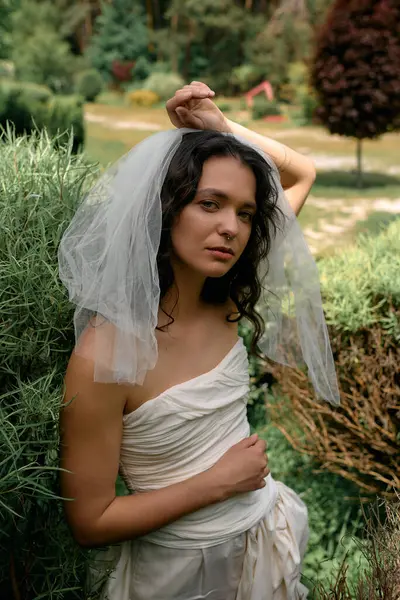 A young bride in a white dress poses gracefully amid lush greenery on a sunny day. — стоковое фото