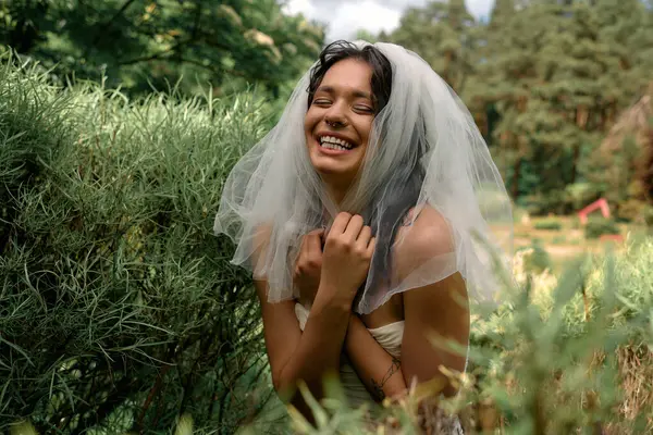 Una joven novia con un vestido blanco sonríe en medio de una exuberante vegetación en un día de verano. - foto de stock
