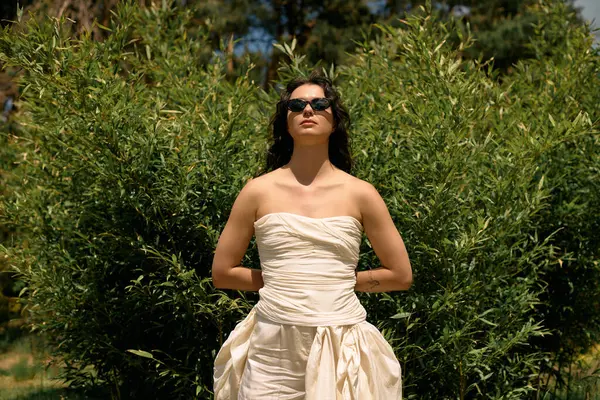 Curly-haired woman stands proudly in a white summer dress surrounded by lush greenery. — стоковое фото
