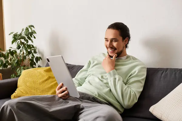 Bello giovane uomo sorride mentre si impegna con il suo tablet in un ambiente confortevole soggiorno. — Foto stock