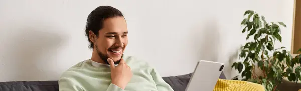 Handsome young man sits comfortably in his living room, smiling as he interacts on his tablet. — Stock Photo