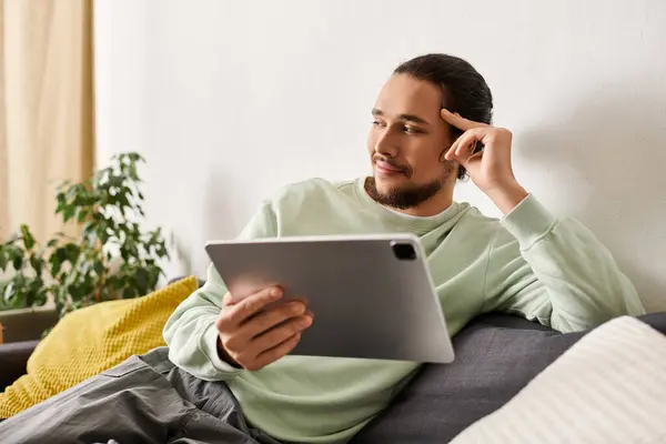 Relaxed young man with a stylish look sits on a couch at home, engaged with a tablet device. — Stock Photo