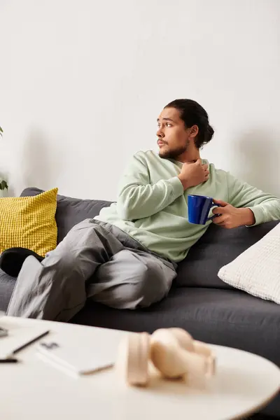 A young handsome man lounges on a sofa, enjoying a cup of warm drink while gazing thoughtfully. — Stock Photo